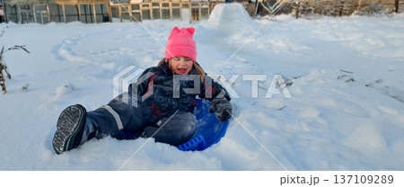 A small child in a pink hat and winter clothes sits on a blue sled in freshly fallen white snow. The child is enjoying playing outside in the cold weather. The child is sliding down a hill on a sled. 137109289
