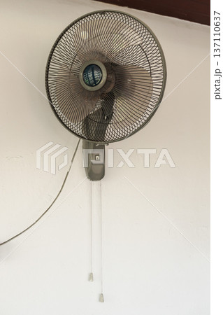 Wall mounted electric fan with pull cords on white wall providing ventilation and cooling in tropical house interior 137110637