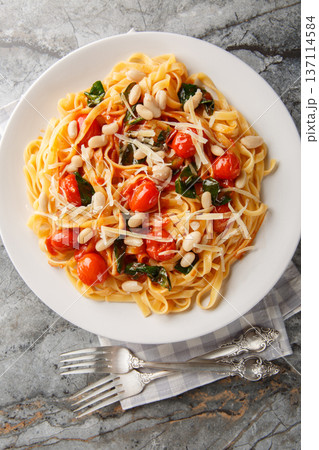 Italian tagliatelle pasta served on a white plate with roasted cherry tomatoes, white beans, spinach and grated parmesan closeup on the plate. Vertical top view 137114584