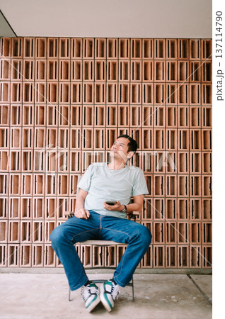Thoughtful Man Holding Phone Sitting by Architectural Terracotta Wall Thoughtful Man Holding Phone Sitting by Architectural Terracotta Wall 137114790