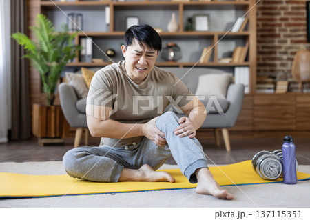 Asian man sitting on yoga mat at home clutching his knee with a pained expression after exercise, depicting knee pain, strain or injury during a workout or stretching session Asian man sitting on yoga mat at home clutching his knee with a pained expression after exercise, depicting knee pain, strain or injury during a workout or stretching session 137115351