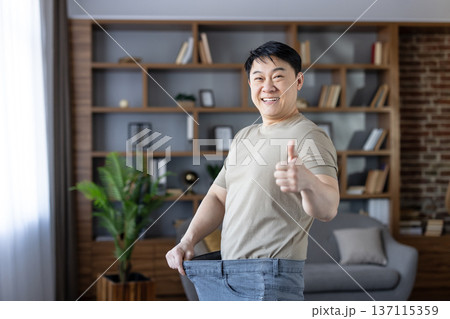 Happy man standing in spacious jeans, giving a thumbs up, celebrating successful weight loss and fitness goals after a healthy transformation journey at home 137115359