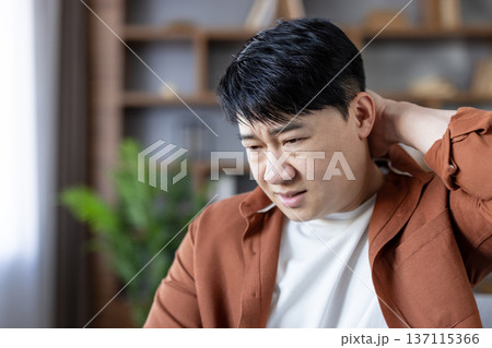 Asian man with a painful expression massaging his stiff neck and shoulder due to discomfort, fatigue, and stress from extended work, signaling a common health problem Asian man with a painful expression massaging his stiff neck and shoulder due to discomfort, fatigue, and stress from extended work, signaling a common health problem 137115366