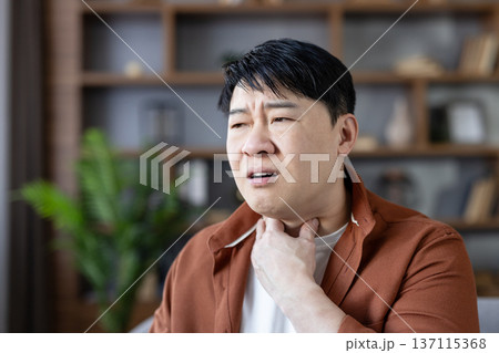 Asian man at home with painful sore throat, touching his neck and grimacing from illness and discomfort, appearing unwell and fatigued while suffering respiratory symptoms 137115368