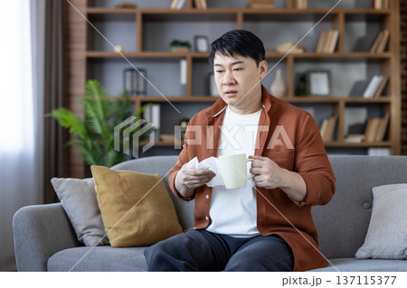 Asian man holding a tissue and a warm drink while sitting on a couch at home, experiencing cold or flu symptoms and feeling sick, needing to rest and recover Asian man holding a tissue and a warm drink while sitting on a couch at home, experiencing cold or flu symptoms and feeling sick, needing to rest and recover 137115377