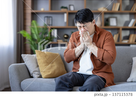 Asian man sitting on a couch at home, suffering from an illness, allergy, or cold, covering his nose with a tissue while sneezing and showing discomfort Asian man sitting on a couch at home, suffering from an illness, allergy, or cold, covering his nose with a tissue while sneezing and showing discomfort 137115378