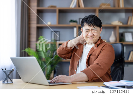 Asian man experiencing severe neck discomfort and pain, rubbing his sore neck while working from home on a laptop, highlighting occupational health issues and poor posture Asian man experiencing severe neck discomfort and pain, rubbing his sore neck while working from home on a laptop, highlighting occupational health issues and poor posture 137115435