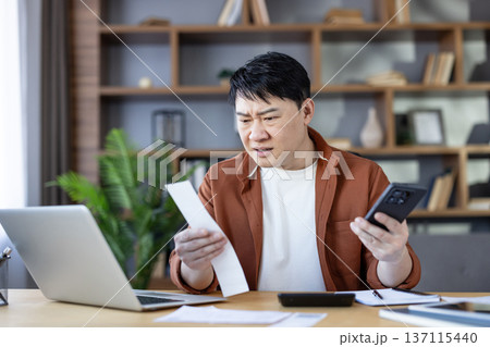 Asian man sitting at a home office desk, holding a receipt and smartphone, looking worried while calculating expenses and managing personal finance or taxes 137115440