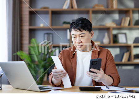 Asian man analyzing a paper receipt and using a modern smartphone for tracking expenses, managing personal finances, or processing invoices while working from his home office desk Asian man analyzing a paper receipt and using a modern smartphone for tracking expenses, managing personal finances, or processing invoices while working from his home office desk 137115442