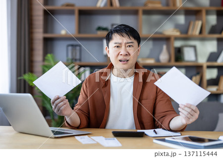 Stressed asian man working from home, holding bills and frustrated with financial problems, calculating expenses, and managing his budget at the office desk 137115444
