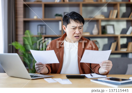 Asian man sitting at a desk, looking bewildered and concerned while holding multiple financial documents, calculating expenses and facing unexpected debts 137115445