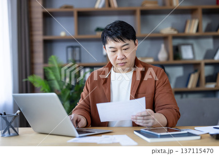 Focused asian man reviewing important documents and bills while using a laptop at his home office desk, diligently managing personal finances and household paperwork Focused asian man reviewing important documents and bills while using a laptop at his home office desk, diligently managing personal finances and household paperwork 137115470