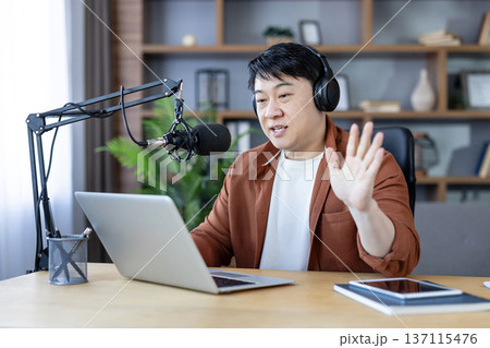 Asian man with headphones speaking into professional microphone, waving hand while recording online podcast or broadcasting live from home studio using a laptop 137115476