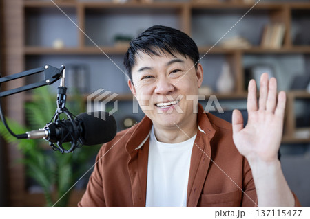 Asian man creating content, smiling and waving, broadcasting a live stream or recording a podcast in a home studio, connecting with his online audience 137115477