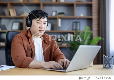 Asian man with headphones concentrating on his laptop, typing and working remotely from a modern home office setting during the day, showing dedication to his online tasks 137115490