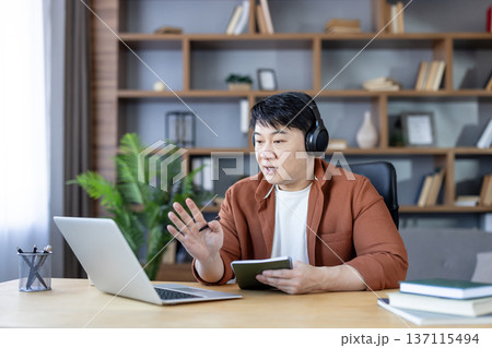 Young asian man wearing headphones, communicating and learning online during a video call on a laptop while working or studying from his modern home office 137115494