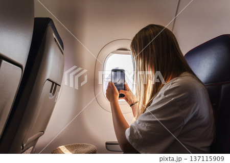Young woman photographing airplane wing through cabin window during flight 137115909