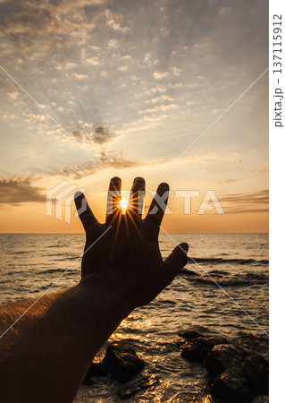 Silhouette hand holding sun over sea horizon at sunset with dramatic sky background 137115912