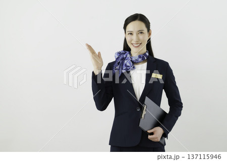 白背景の紺のスーツにリボン、ネームタグをつけて手をかざす笑顔の日本人女性　コンシェルジェ 137115946
