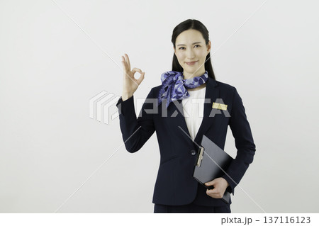 白背景の紺のスーツにリボン、ネームタグをつけて指で丸のokのポーズをする笑顔の日本人女性　コンシェル 137116123