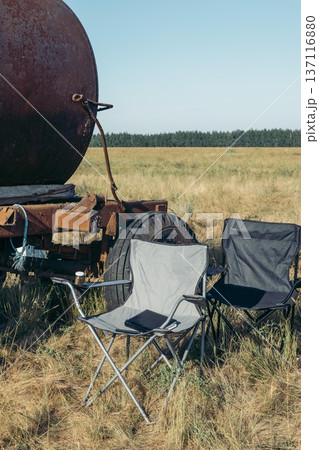 Two folding chairs sit in a field near a rusty agricultural tank. The landscape is open with tall grass and a clear blue sky. Two folding chairs sit in a field near a rusty agricultural tank. The landscape is open with tall grass and a clear blue sky. 137116880