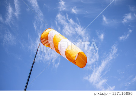 orange and white striped windsock indicating wind direction on pole against a clear blue sky with light clouds, concept of weather monitoring, aviation, airfield safety orange and white striped windsock indicating wind direction on pole against a clear blue sky with light clouds, concept of weather monitoring, aviation, airfield safety 137116886