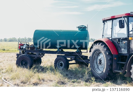 red agricultural tractor parked on rural field with water tank trailer under blue sky open countryside landscape with large copy space banner composition concept of agriculture, farming equipment 137116890