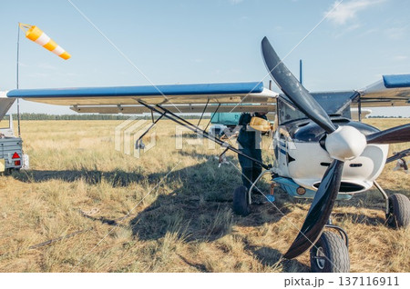 man refueling small white aircraft on grassy airfield, aviation worker in green uniform during sunset, rural field, aviation service, pilot training, countryside tourism 137116911