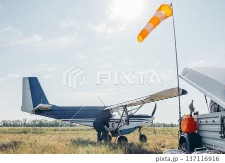 man refueling small blue aircraft on grassy airfield, aviation worker in green uniform during sunset, rural field, aviation service, pilot training, countryside tourism 137116916