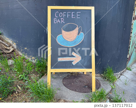 Chalkboard sign with drawing of coffee cup and arrow standing on street near wall and grass outdoors. Cafe direction sign, street advertising, urban cafe culture and small business navigation 137117012