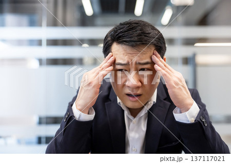 Asian businessman experiencing a severe headache and high stress while working in a modern office, pressing temples to relieve the pain and showing a frustrated expression 137117021
