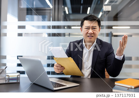 Asian businessman receiving a layoff letter from an envelope at his desk, expressing extreme frustration and disbelief about the bad news in a modern office Asian businessman receiving a layoff letter from an envelope at his desk, expressing extreme frustration and disbelief about the bad news in a modern office 137117031