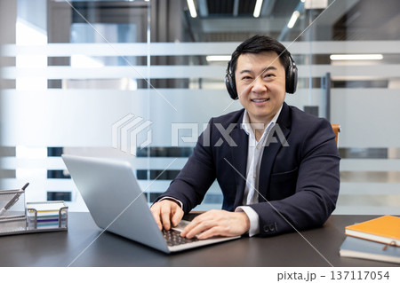 Asian businessman smiling while wearing headphones and typing on a laptop, working at a modern office desk with a transparent partition in the background Asian businessman smiling while wearing headphones and typing on a laptop, working at a modern office desk with a transparent partition in the background 137117054