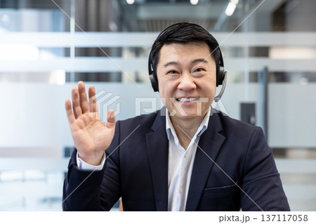 Asian businessman wearing a headset and smiling. Waving his hand hello or goodbye during a remote video conference from a modern office setting. Representing online communication and customer service 137117058