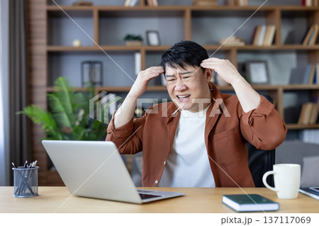 Overworked asian man clutching his head, in pain and frustration from a severe headache or migraine while working on a laptop at a cluttered home office desk Overworked asian man clutching his head, in pain and frustration from a severe headache or migraine while working on a laptop at a cluttered home office desk 137117069