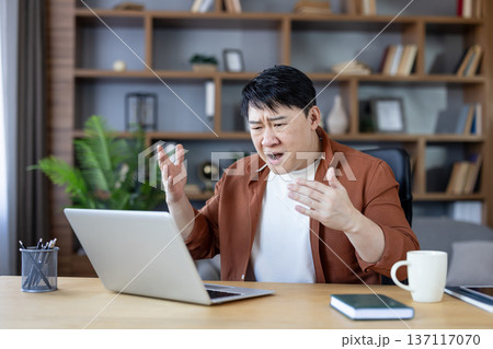 Asian man frustrated and angry during a video call on laptop, showing stress and technical connection problems while working from his home office during remote work session 137117070