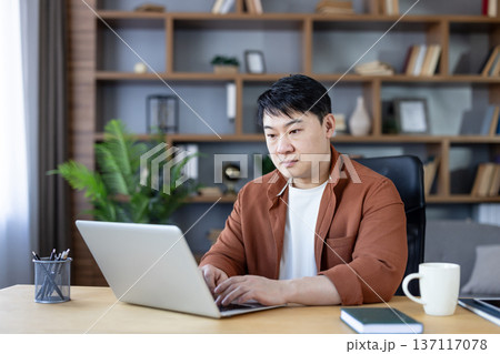Asian man focused on laptop in a modern home office, working remotely on freelance business tasks at his desk, typing, researching and planning a project 137117078