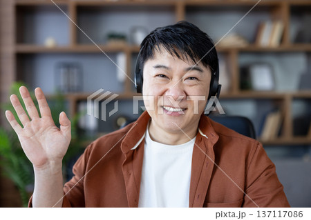 Smiling asian man in headset waving during an online video call, greeting colleagues or students from a modern home office while working or teaching remotely via webcam Smiling asian man in headset waving during an online video call, greeting colleagues or students from a modern home office while working or teaching remotely via webcam 137117086