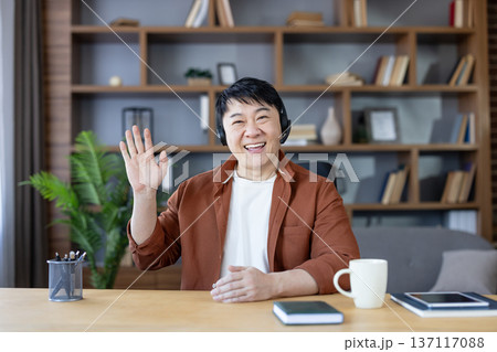 Smiling asian man wearing a headset and brown shirt, sitting at a desk in a home office, looking at the camera and waving during a virtual meeting, video conference, or online communication 137117088