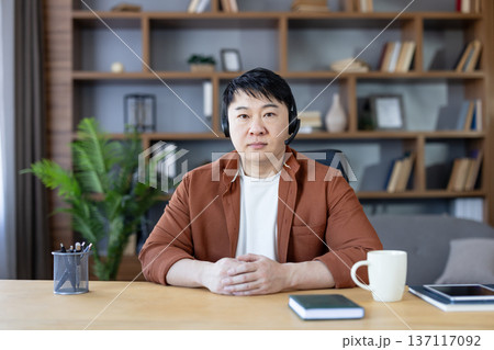 Asian man in a home office wearing a headset, focused on a video call or webinar with hands clasped on the desk beside laptop, notebook and mug Asian man in a home office wearing a headset, focused on a video call or webinar with hands clasped on the desk beside laptop, notebook and mug 137117092