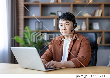Asian man wearing a headset and typing on a laptop, engaged in remote work or online meeting, leveraging technology for communication from a home office environment Asian man wearing a headset and typing on a laptop, engaged in remote work or online meeting, leveraging technology for communication from a home office environment 137117110