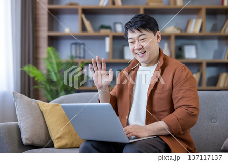 Asian man smiling, waving hand, and communicating during a virtual meeting on his laptop, sitting comfortably on a gray sofa in a modern home living room 137117137