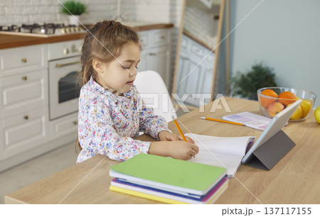 Busy child making homework with tablet and paper notebook, learning and sitting at kitchen desk 137117155