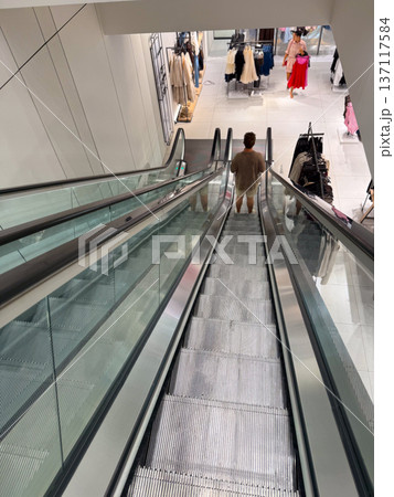 A person descends on a glass escalator inside a modern shopping mall retail space. Consumer movement, urban lifestyle, shopping experience and architectural design within commercial culture. A person descends on a glass escalator inside a modern shopping mall retail space. Consumer movement, urban lifestyle, shopping experience and architectural design within commercial culture. 137117584