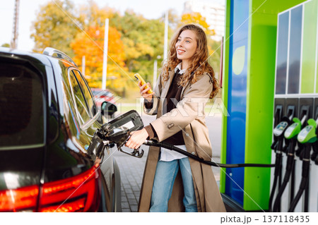 Beautiful woman filling car with gasoline at gas station. Online pay. Woman using app on phone. Beautiful woman filling car with gasoline at gas station. Online pay. Woman using app on phone. 137118435