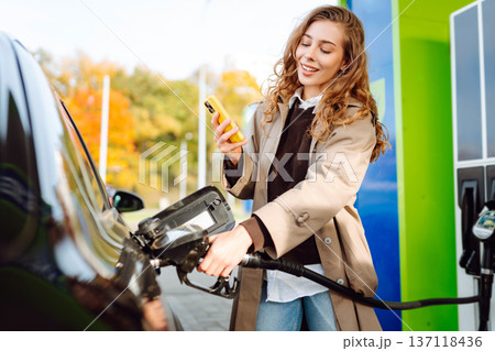 Beautiful woman filling car with gasoline at gas station. Online pay. Woman using app on phone. Beautiful woman filling car with gasoline at gas station. Online pay. Woman using app on phone. 137118436