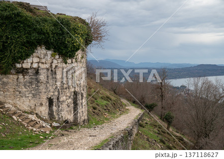Stone fortress wall with ivy on hillside path at Anacopia fortress in Abkhazia overlooking Black Sea under cloudy sky, Historic architecture and coastal landscape suitable for tourism marketing 137118627