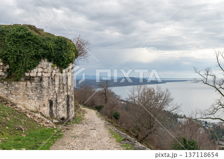 Stone fortress wall with ivy on hillside path at Anacopia fortress in Abkhazia overlooking Black Sea under cloudy sky, Historic architecture and coastal landscape suitable for tourism marketing 137118654