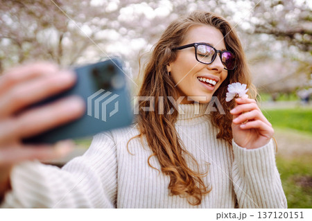 Young woman takes selfie against background of flowering trees in park. Concept of relaxation, fun. Young woman takes selfie against background of flowering trees in park. Concept of relaxation, fun. 137120151