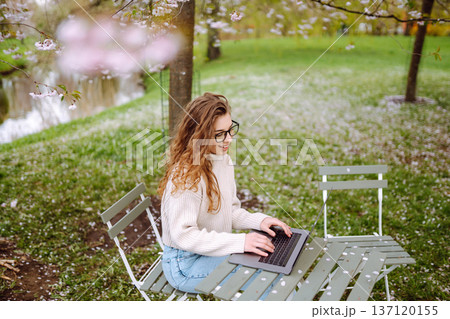 A young woman at a table in a blooming park works with a laptop. Freelancing, weekend concept. 137120155